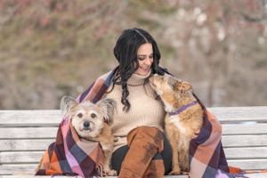 Woman wrapped in plaid blanket sitting on bench with two dogs in autumn park. Pet Photo by Xenophon Photography in Maine.