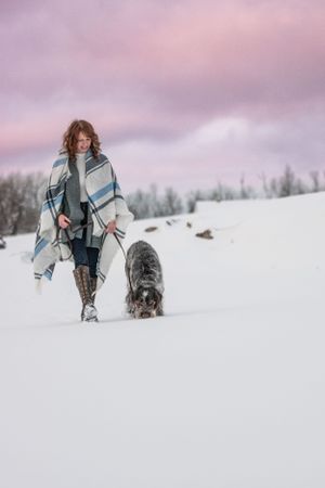 Woman walking her Wirehaired Pointing Griffon dog through deep snow under a pink winter sunset sky. Canine Photographer in Maine