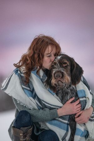 Red-haired woman wrapped in a plaid blanket kissing her Wirehaired Pointing Griffon dog in a snowy winter landscape. Pet Photographer