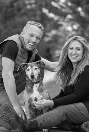 Black and white photo of a smiling couple posing with their senior mixed breed dog outdoors. Pet Photographer in Maine