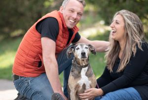 Laughing couple in casual outdoor wear sitting with their senior brindle mixed breed dog in a park. Pet Family Photographer in Maine