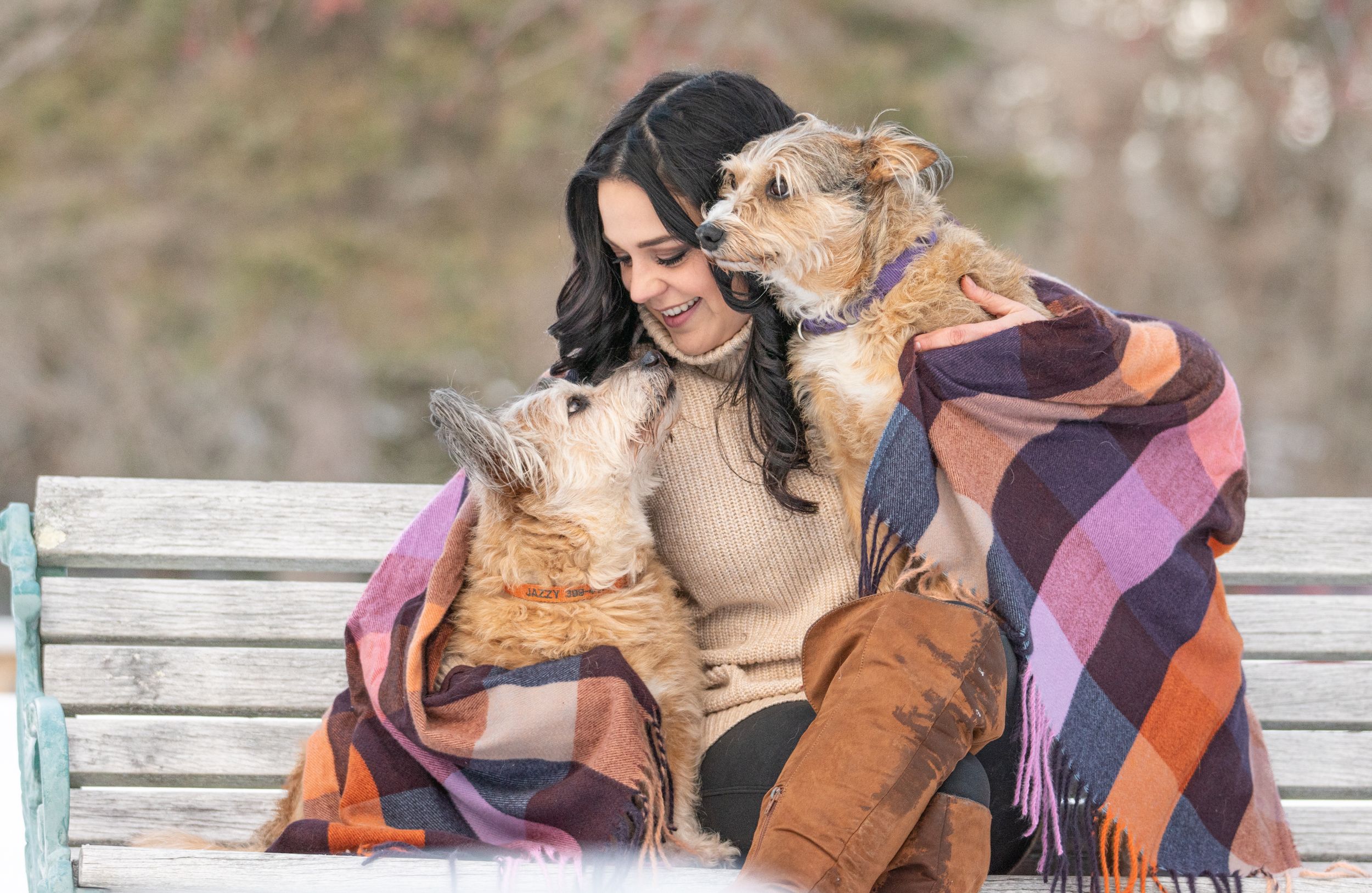 Smiling woman on bench cuddling two dogs under a colorful plaid blanket outdoors. Pet Photo by Xenophon Photography in Maine.