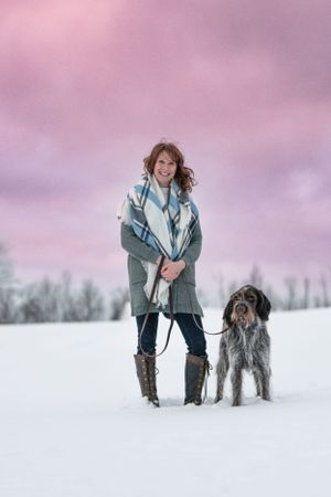 Woman standing with her Wirehaired Pointing Griffon dog on a snowy field under a pink cloudy sky. Dog Photographer in Maine