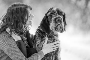 Woman gently holding her Wirehaired Pointing Griffon dog in a tender black and white outdoor portrait. Canine Photographer in Maine