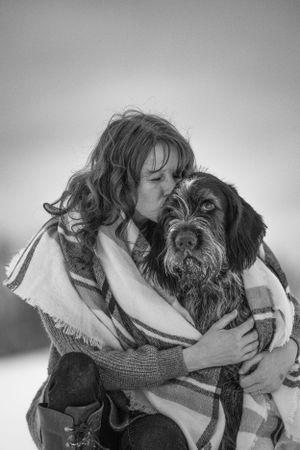 Woman kissing her Wirehaired Pointing Griffon dog wrapped in plaid blanket in snowy winter landscape, black and white photo. Pet Photography