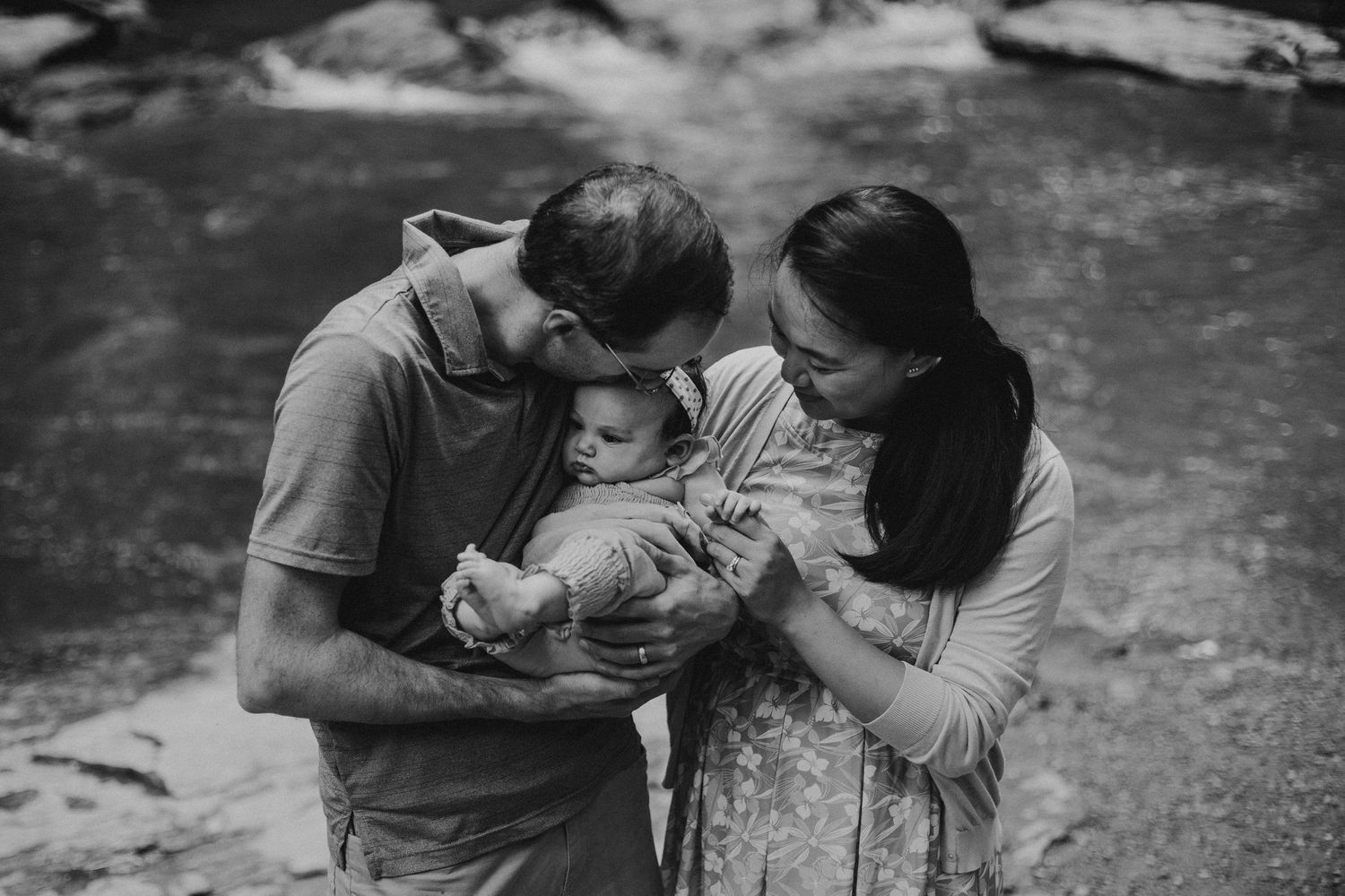 black & white photo looks down at two parents who are hugging their baby; there is a stream in the background