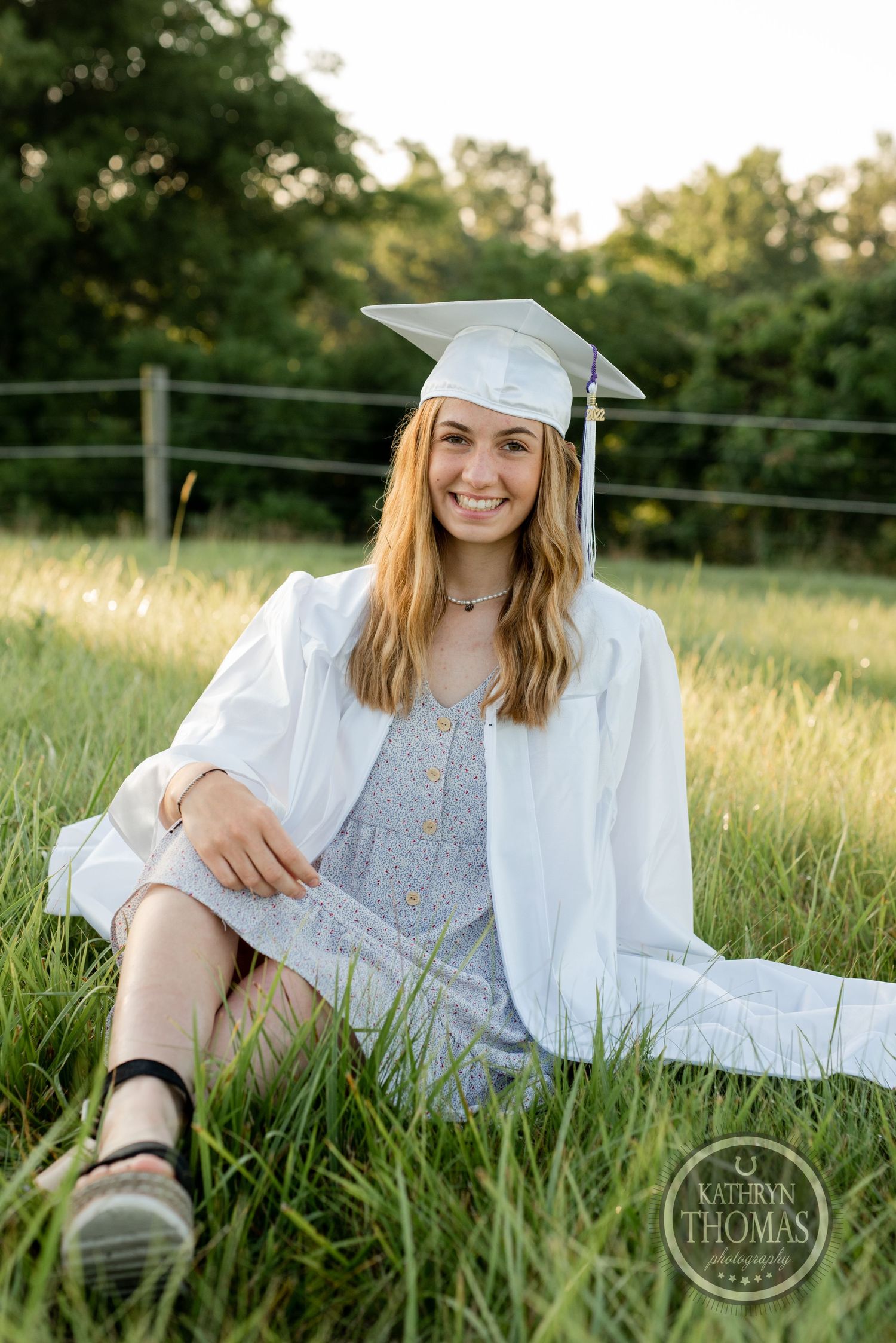 Graduate in white gown and grey dress relaxes in grassy field during golden hour photo session.