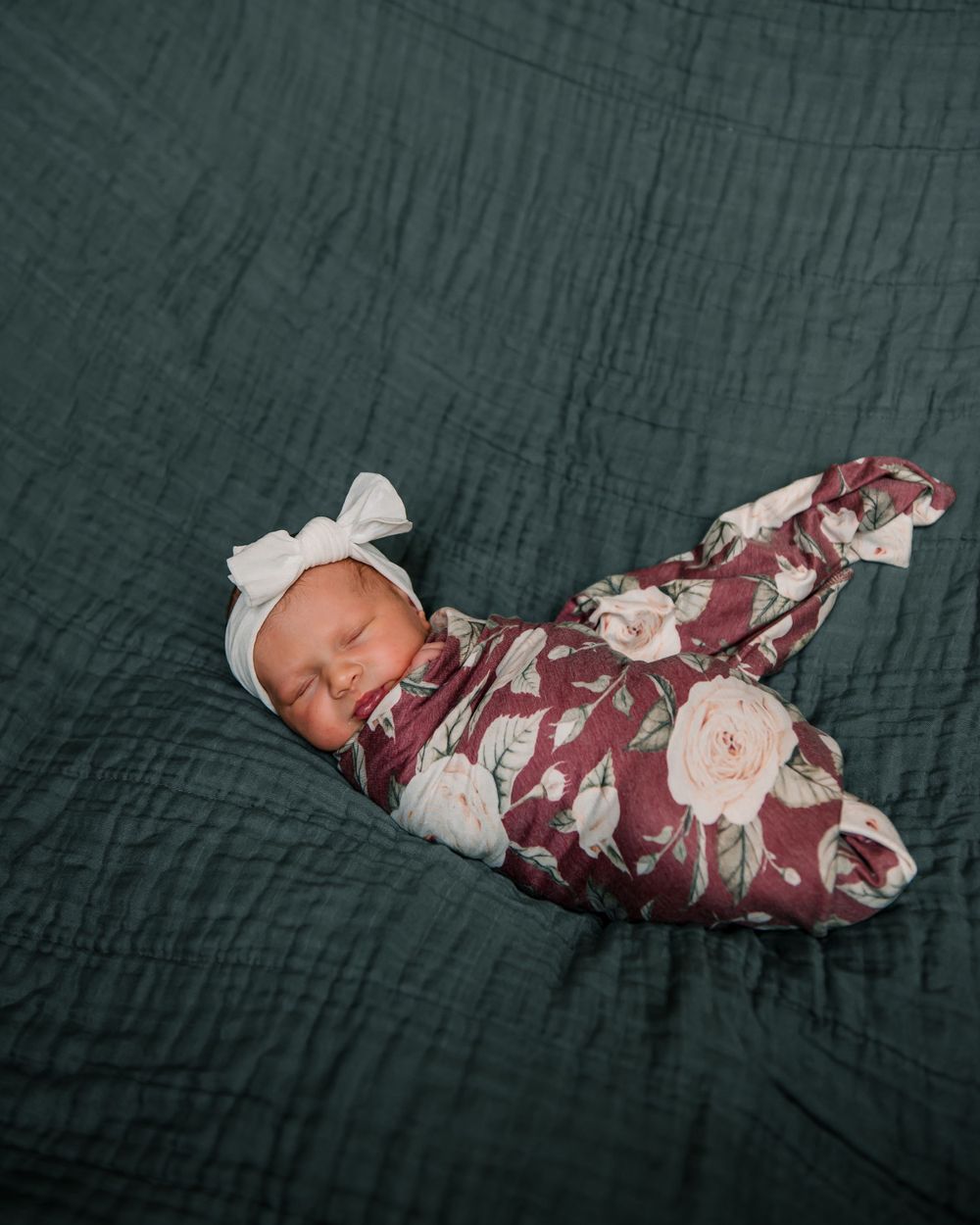 Sleeping newborn wrapped in floral swaddle on dark green textured bedding.