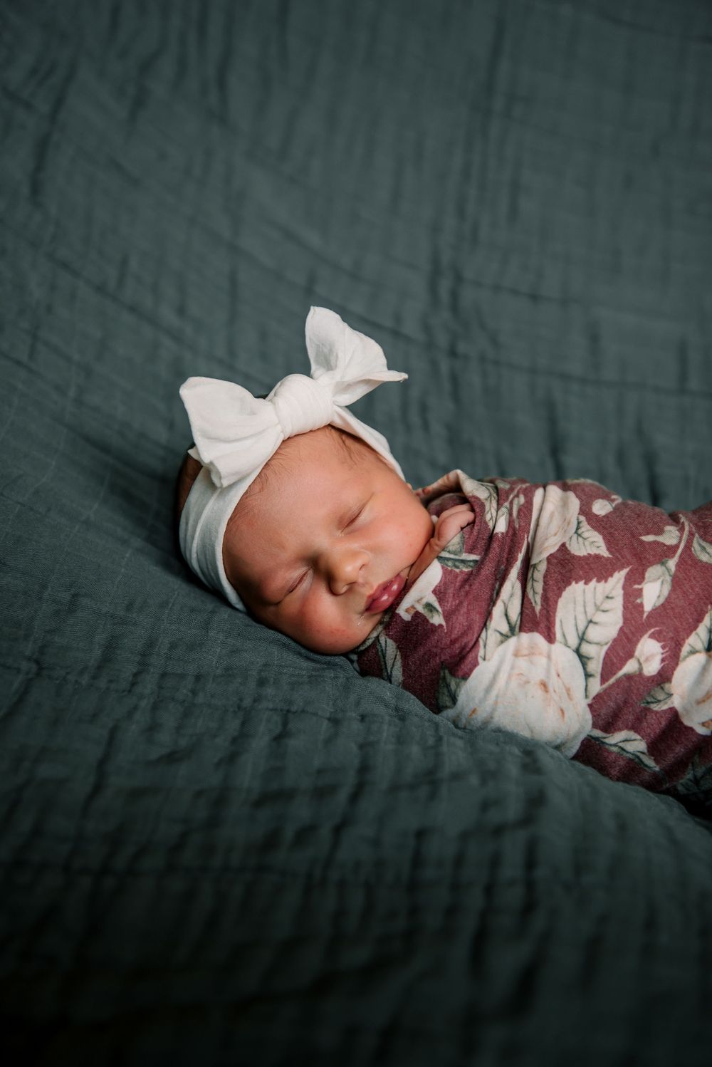 Peaceful newborn resting on dark green bedding wearing floral wrap and white bow.