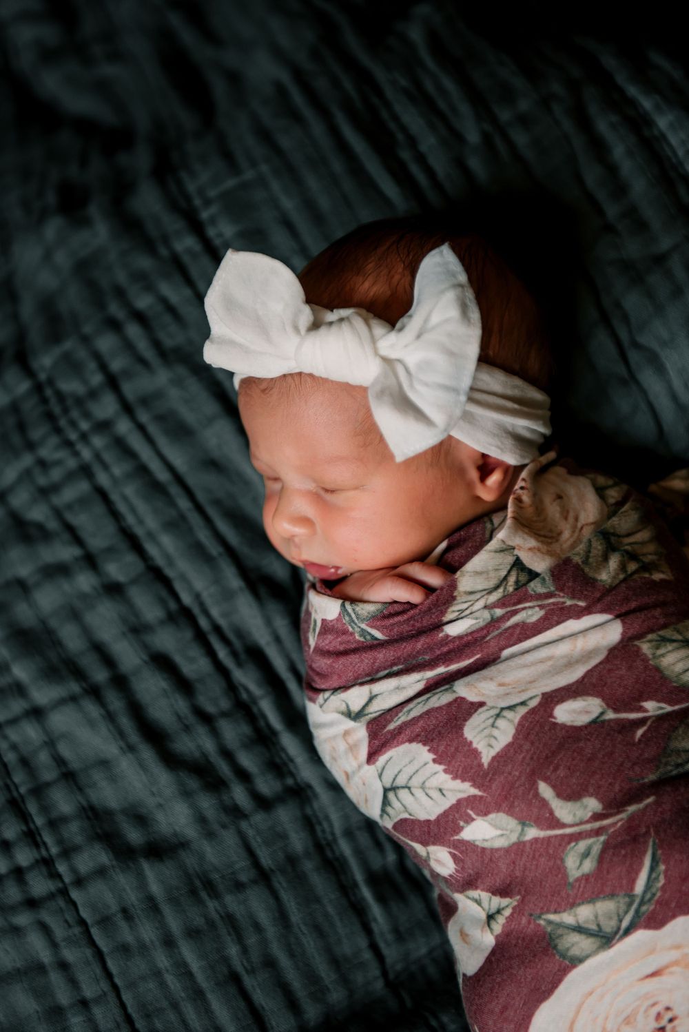 Close-up of sleeping newborn with white bow headband on dark textured fabric.