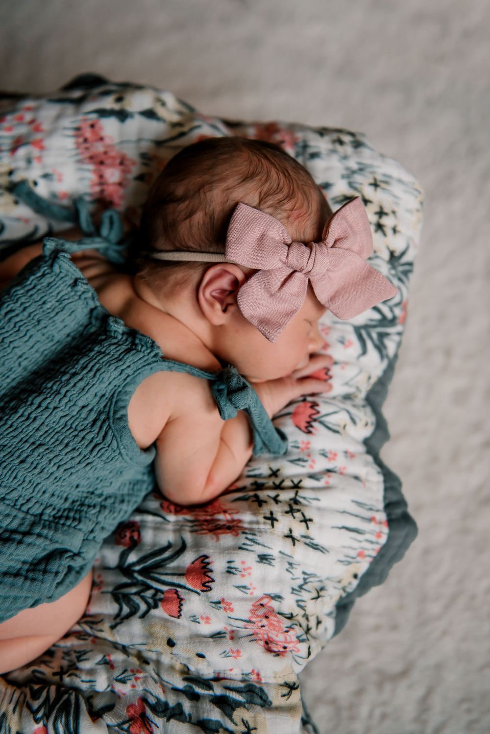 Sleeping newborn with pink bow wearing teal swaddle on floral bedding.