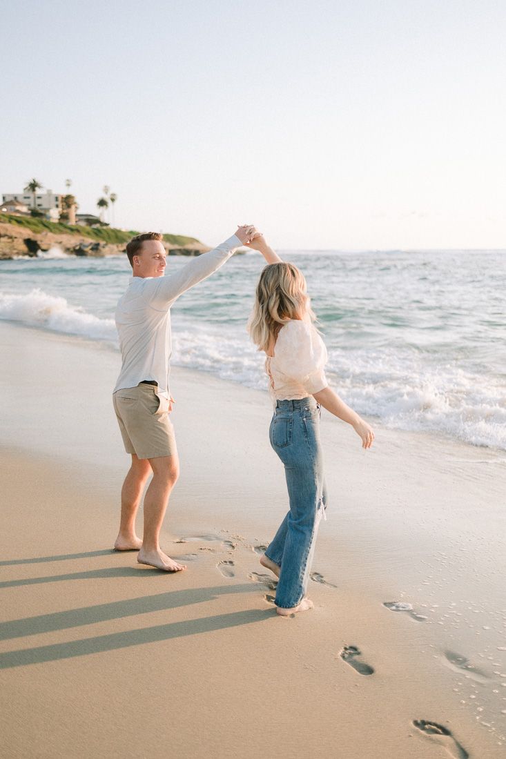 Couple dancing together on a sunny beach with waves and coastal cliffs in the background.