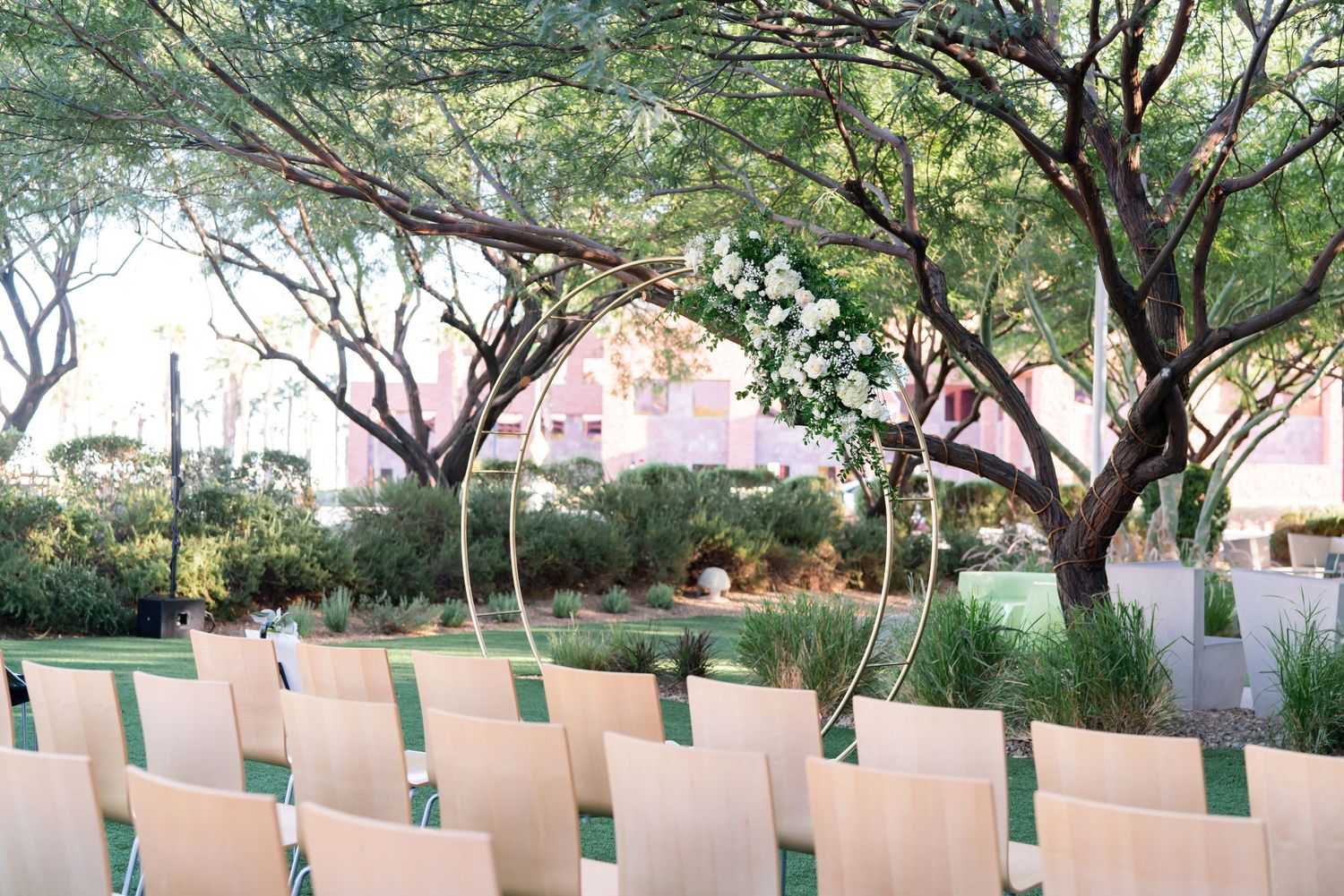 Outdoor wedding ceremony setup with rows of chairs under desert trees and floral arch.