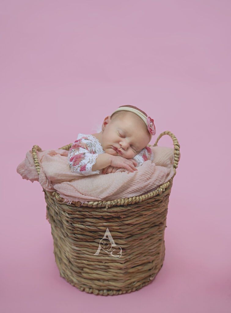Sleeping baby nestled in wicker basket with pink blanket against rosy background.