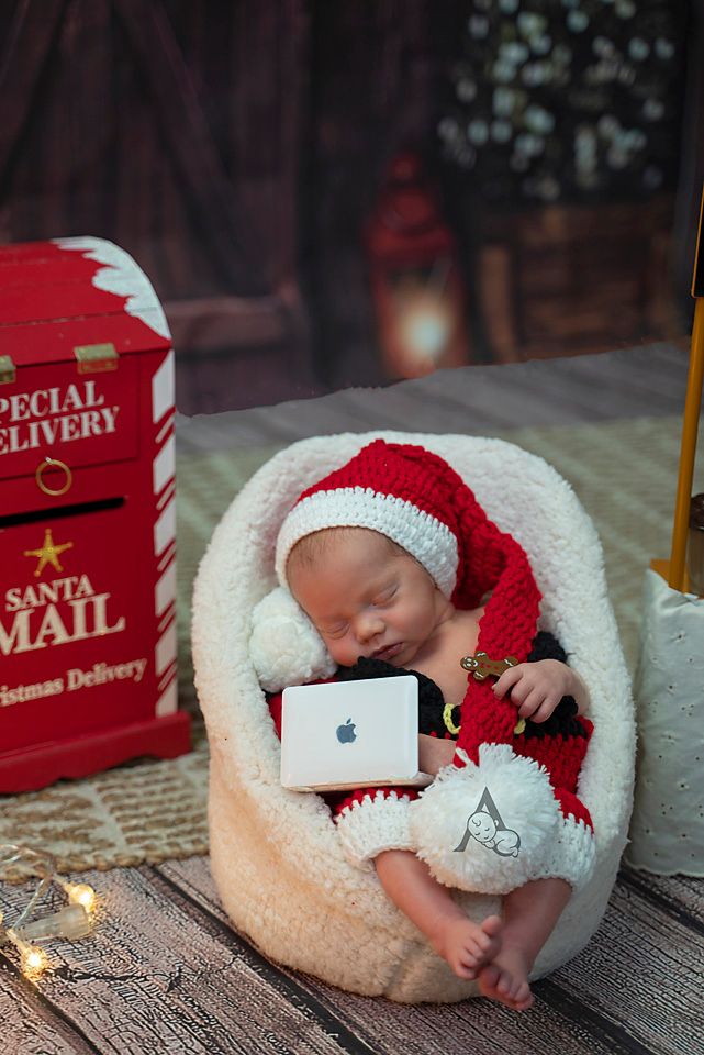 Newborn in red Santa hat sleeps with miniature laptop next to red Christmas mailbox decoration.