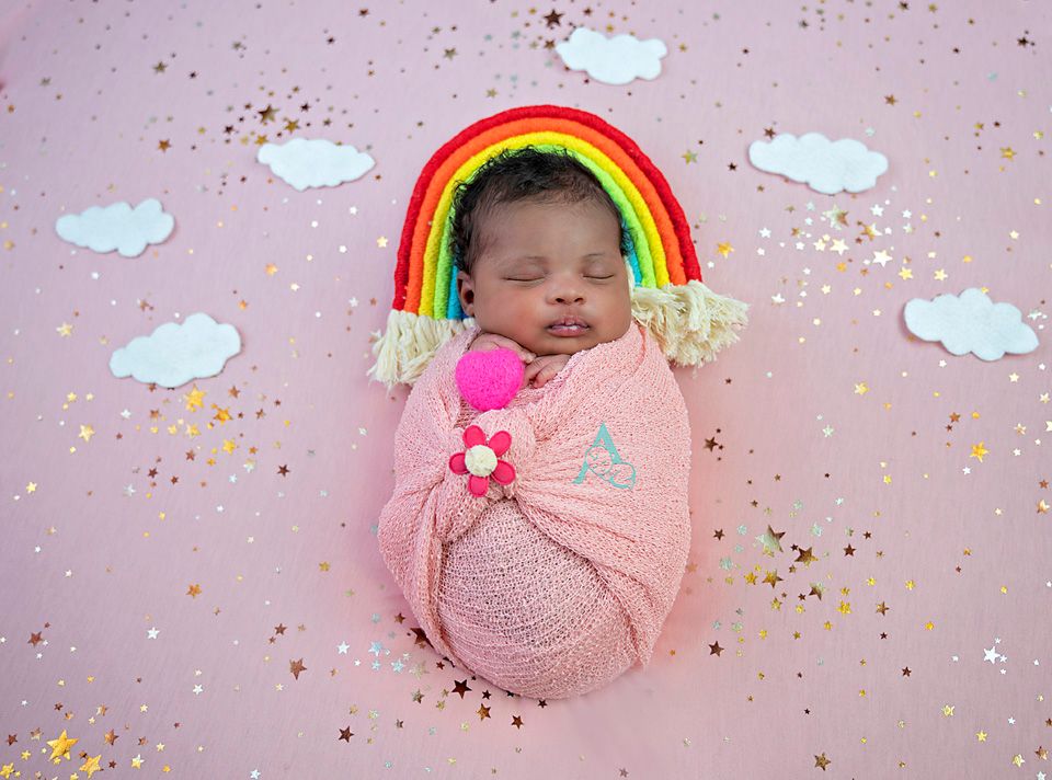 Sleeping newborn wrapped in pink blanket under decorative rainbow and clouds on pink background.