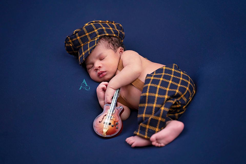Sleeping newborn in plaid outfit holding miniature guitar on blue background.