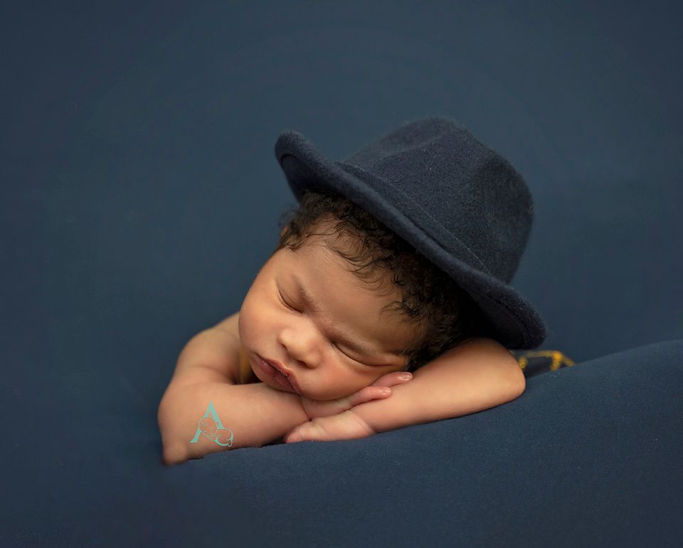 Sleeping newborn wearing dark fedora hat on navy blue backdrop.