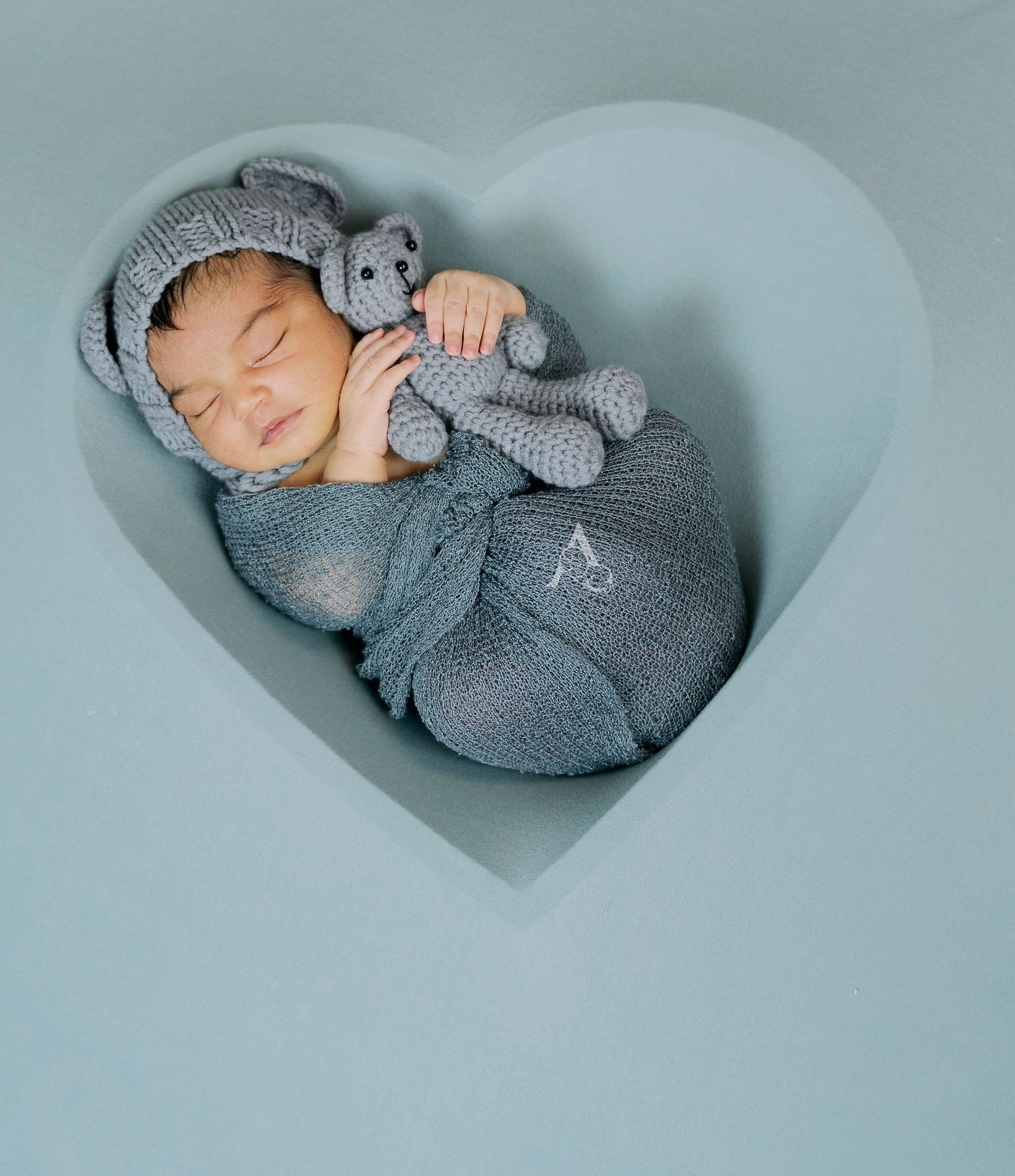 Newborn wrapped in grey swaddle sleeping with matching stuffed toy in heart-shaped frame.