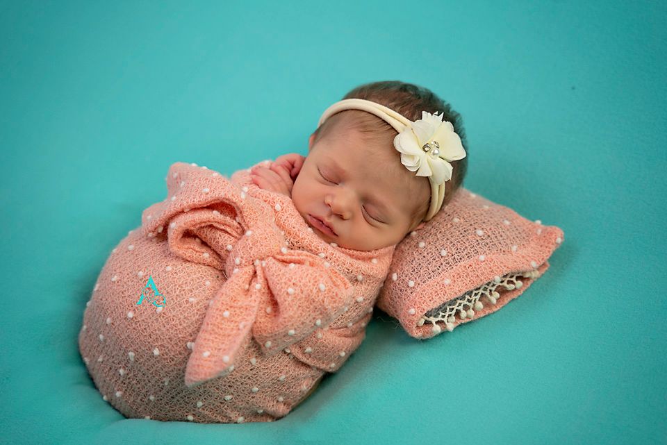 A sleeping newborn swaddled in a peach-colored wrap with pearls and wearing a cream flower headband on turquoise backdrop.