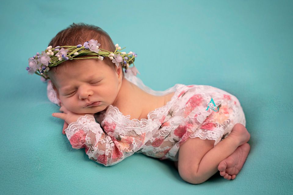 A sleeping newborn in a pink floral dress and floral crown rests on a turquoise background.