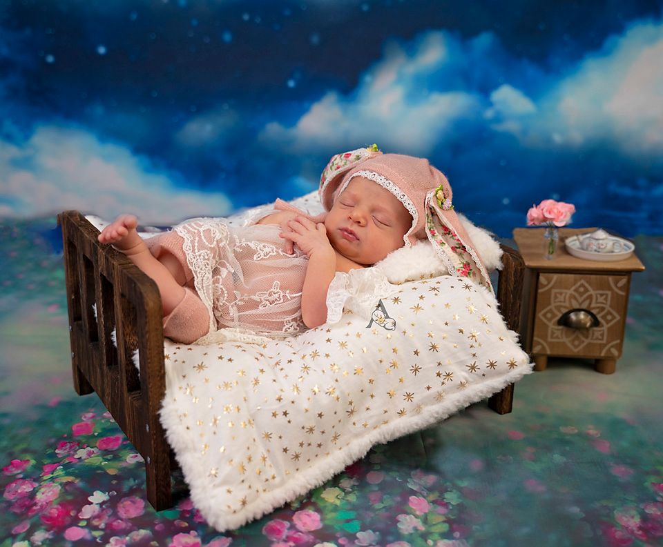 A newborn sleeps in a wooden bed with sparkly bedding and antique nightstand against a starry background.