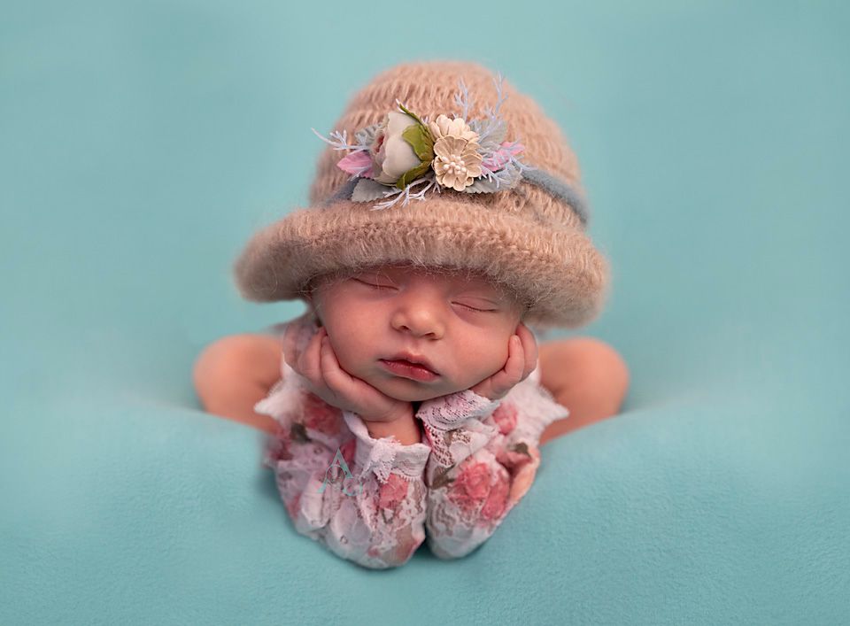 A newborn wearing a pink knit hat with flowers poses against a turquoise background.