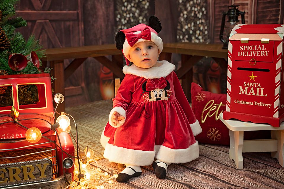 Baby dressed in red Santa dress outfit next to vintage mailbox and Christmas decor.