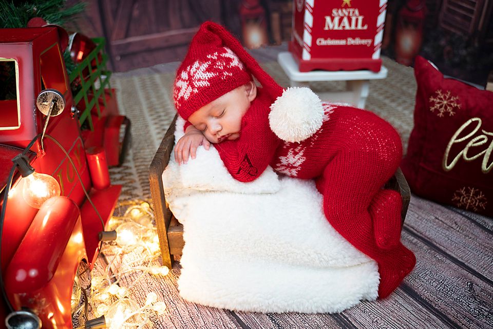 Sleeping baby in red Christmas outfit on white pillow next to decorative mailbox.