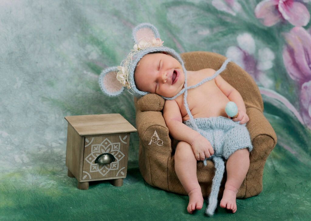 Baby wearing mouse ears resting on miniature armchair with vintage side table.