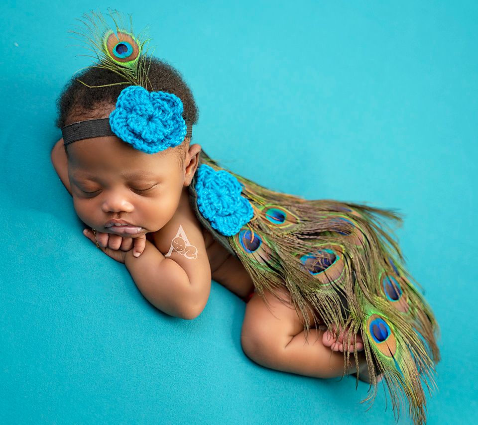 Artistic newborn portrait with peacock feathers and blue flowers on turquoise background.