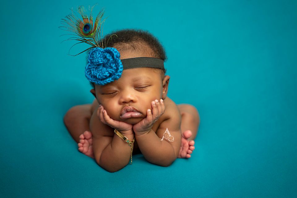 Newborn photo with peacock feather and blue flower headband against turquoise backdrop.