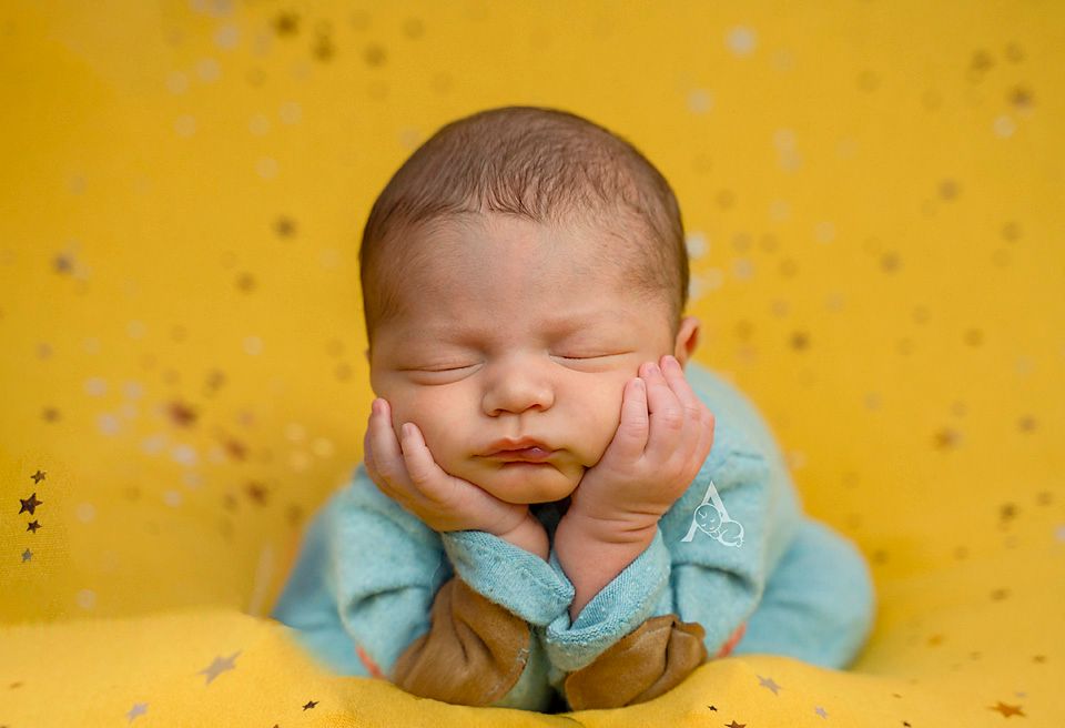 Peaceful napping newborn wearing turquoise outfit against bright yellow backdrop.