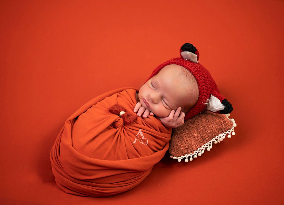 Newborn wrapped in red fabric with fox hat on rustic brown background.