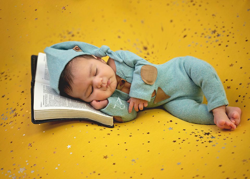 Sleeping newborn in teal outfit resting on open book with glittery yellow background.
