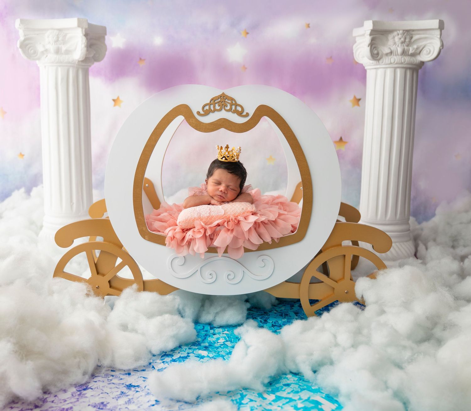 Dreamy princess-themed newborn photography setup with white columns and golden carriage against cloudy backdrop.