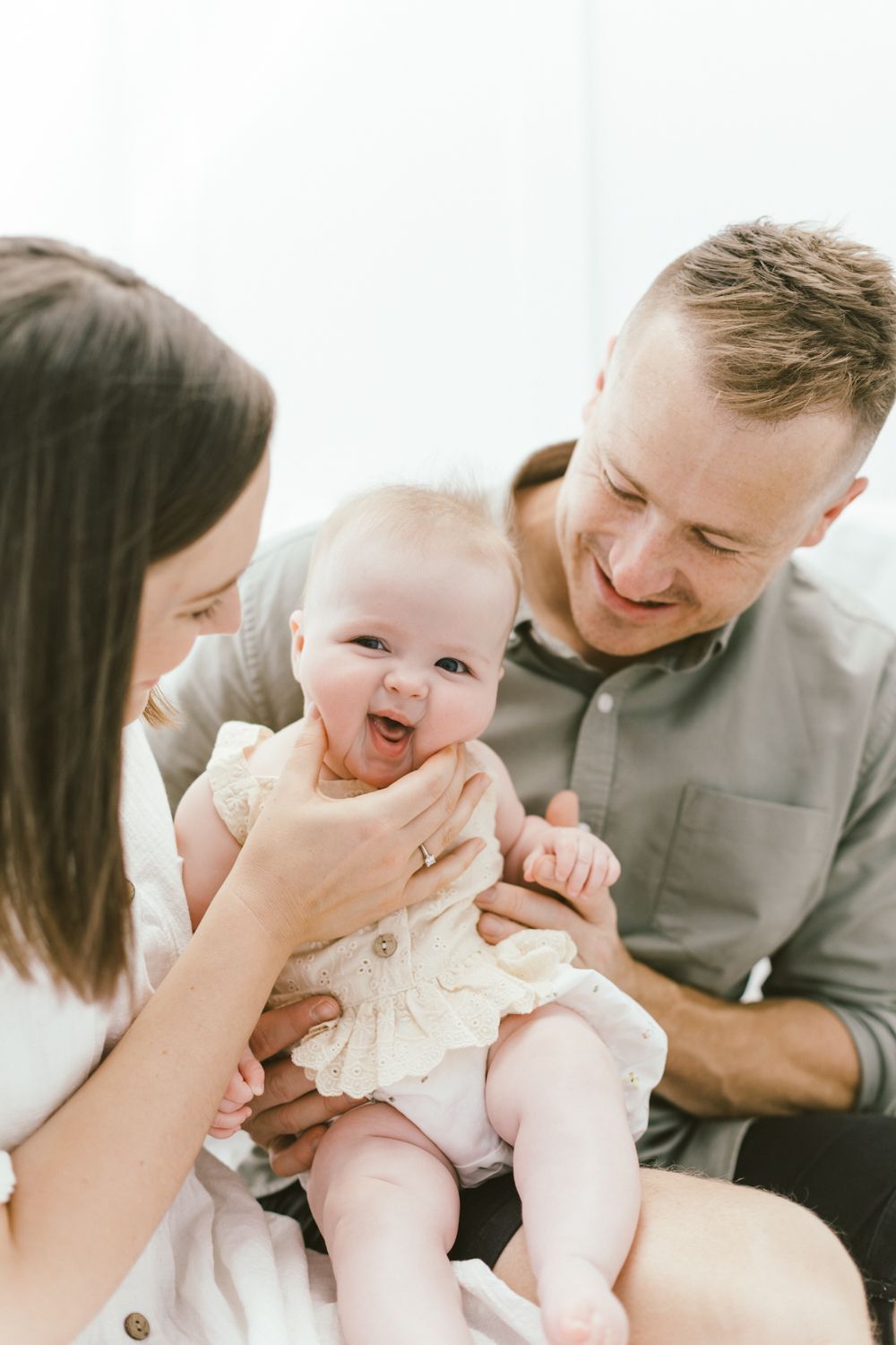 Happy baby in cream dress laughs while being held by parents against white backdrop.
