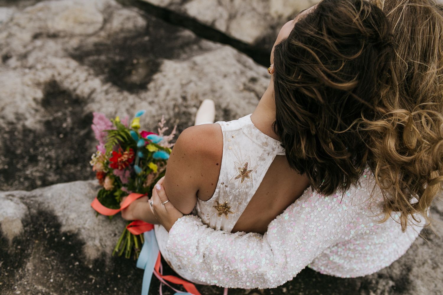 Two brides embracing with colorful bouquet on rocky outdoor setting.
