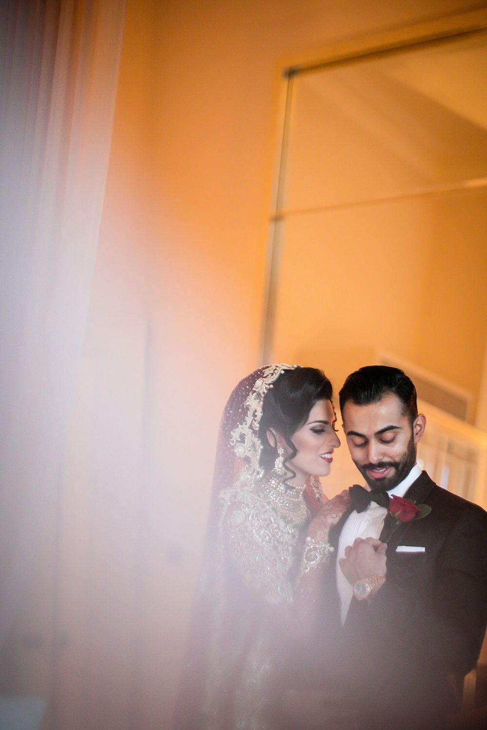 An intimate moment captured through sheer curtains shows a couple in elegant wedding attire.