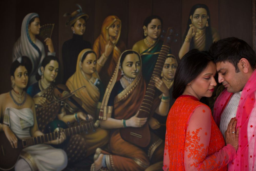 A couple in vibrant attire poses intimately against a backdrop of classical Indian musical artwork.