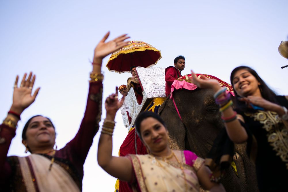 People celebrate joyfully outdoors, raising their hands and traditional decorative items against a blue sky.