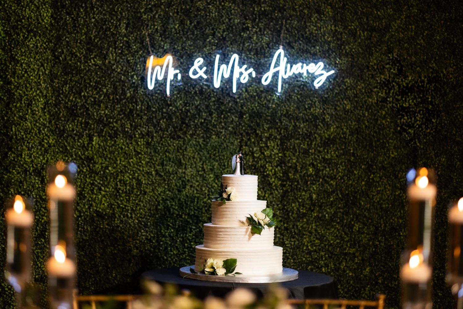 Elegant wedding reception decor with illuminated name sign and candlelight against green hedge wall.