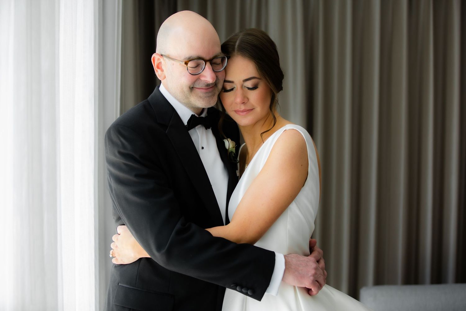 Tender embrace between couple during wedding day in light-filled room with drapes.