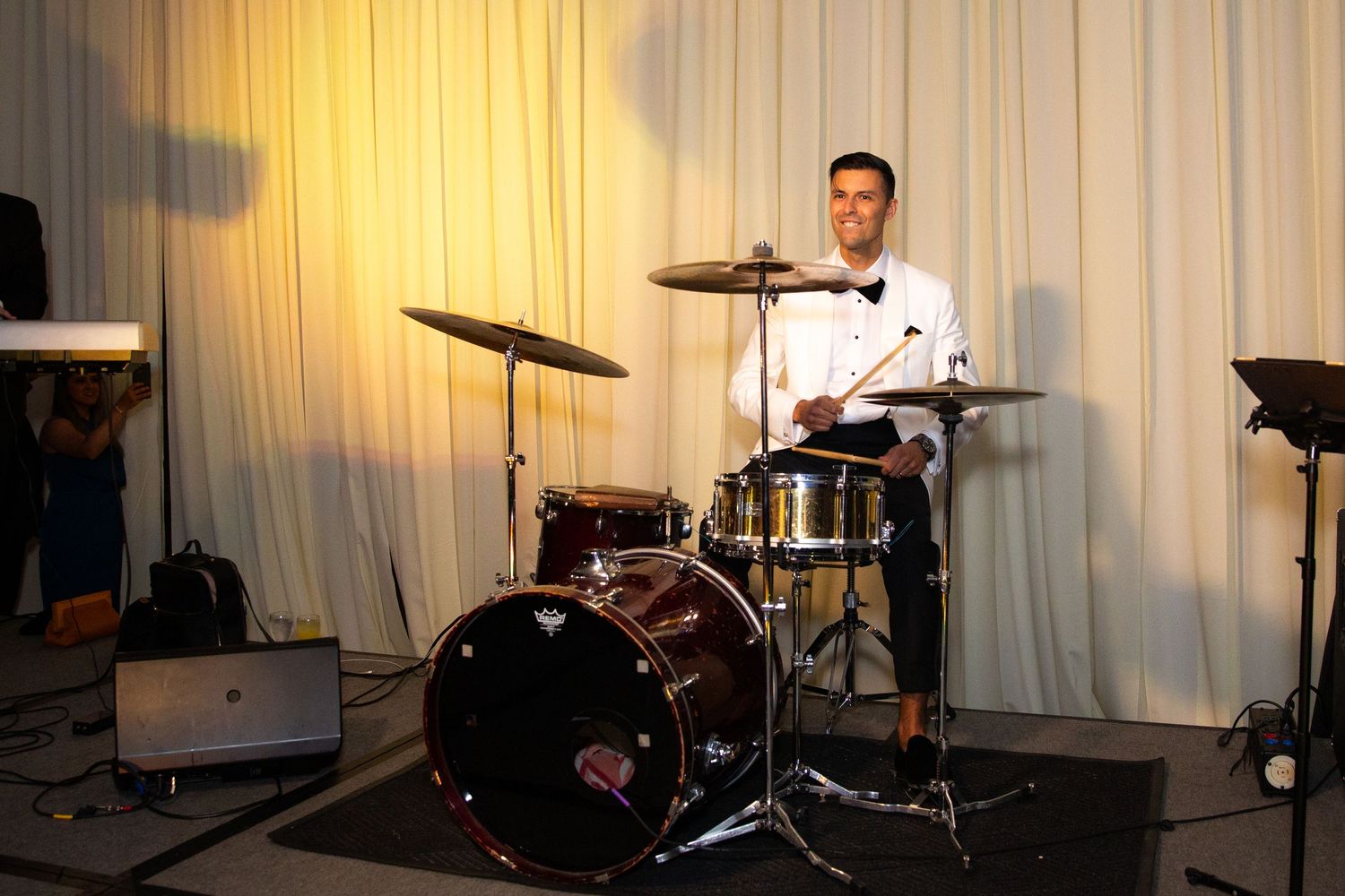 Musicians perform at a wedding reception with drums and instruments against an ivory curtain backdrop.