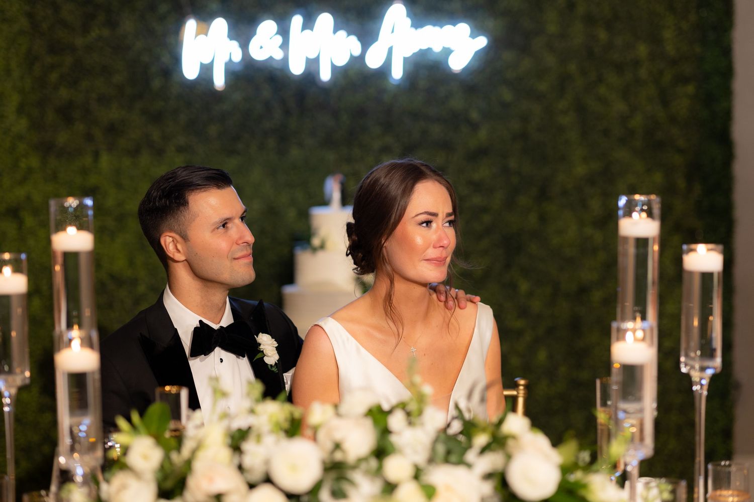 Wedding reception head table with elegant lighting and greenery backdrop.