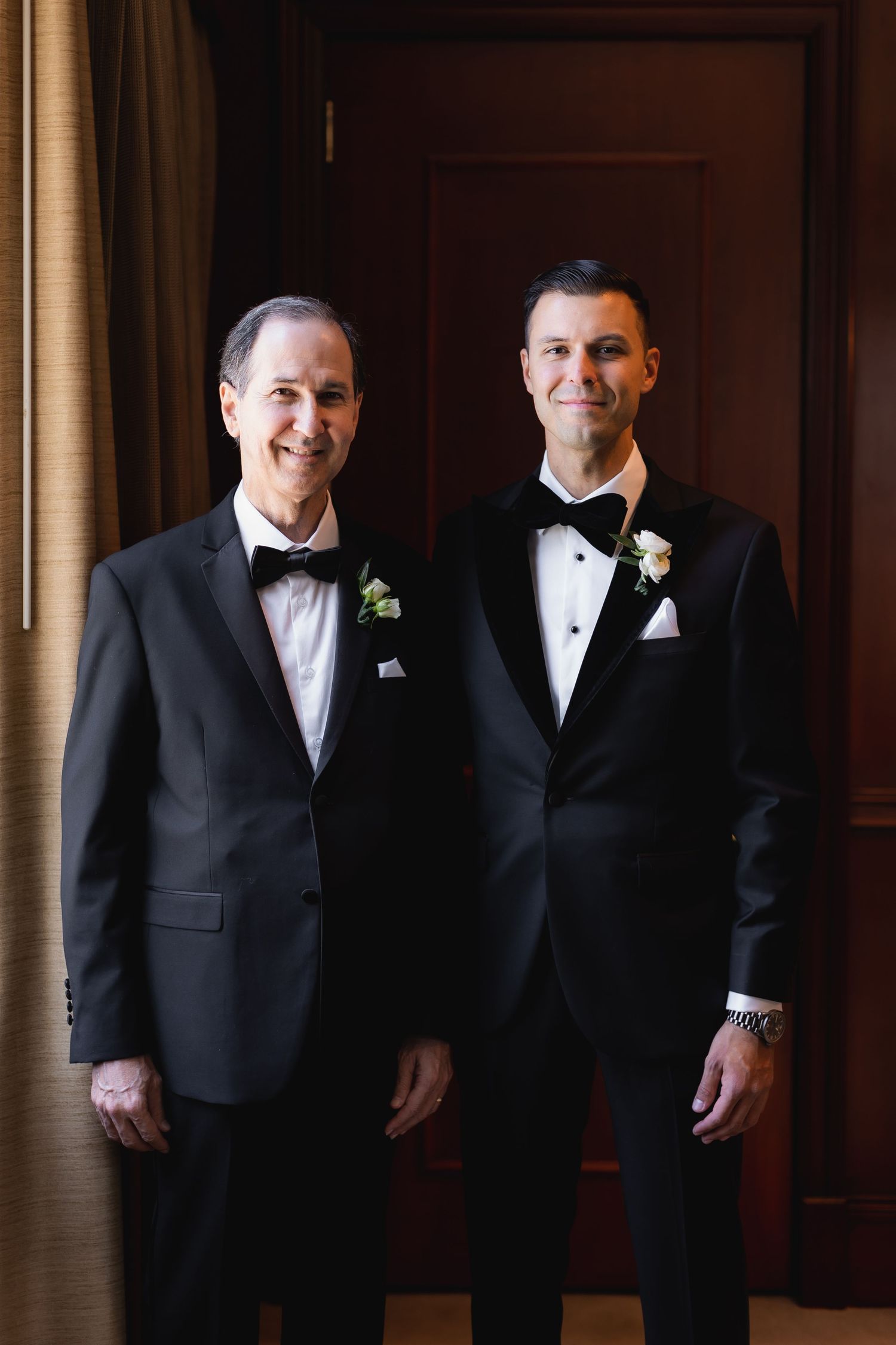 Groomsmen in black tuxedos stand together against wooden paneled walls.