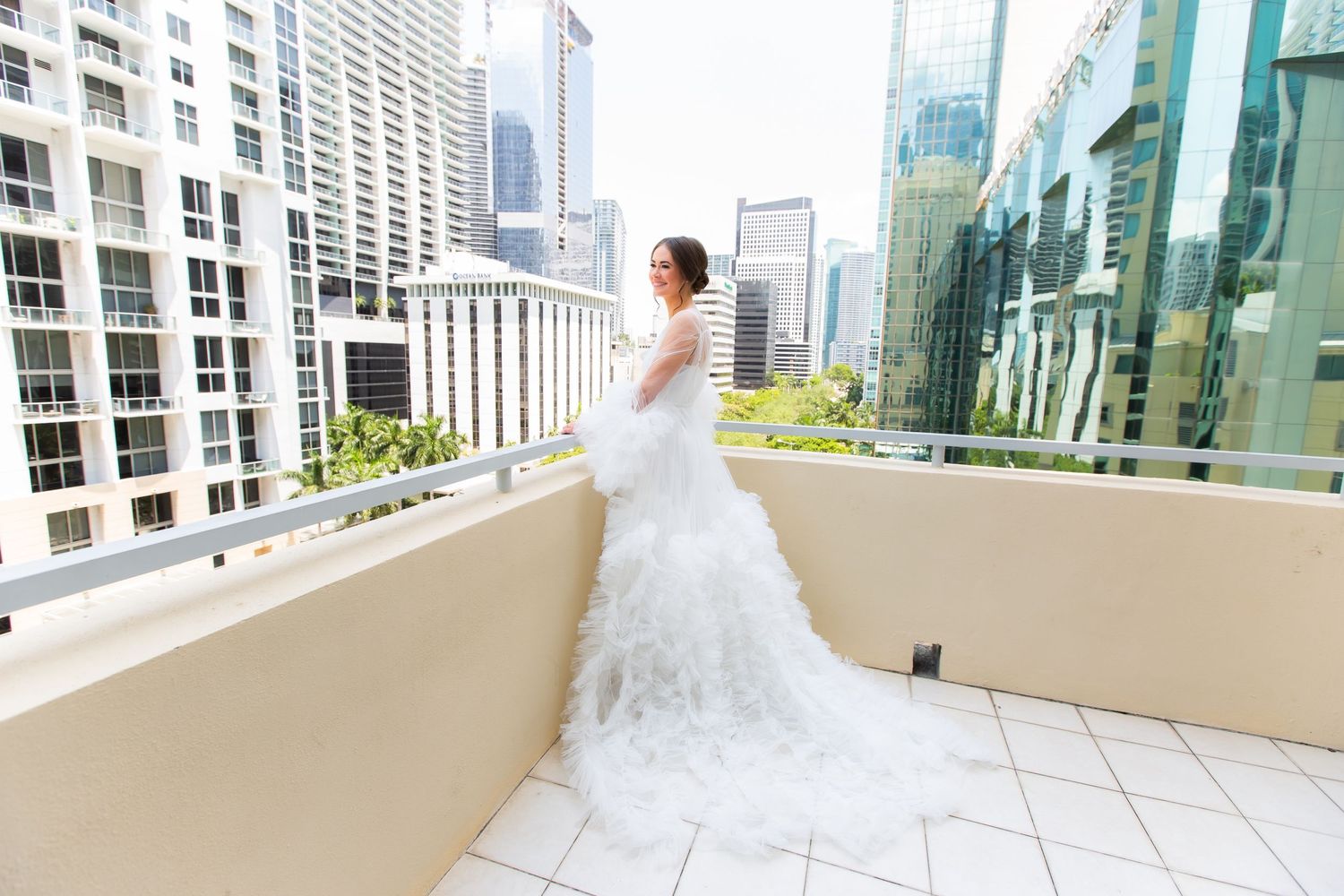 A bride in white poses on a modern urban balcony with city skyscrapers in the background.