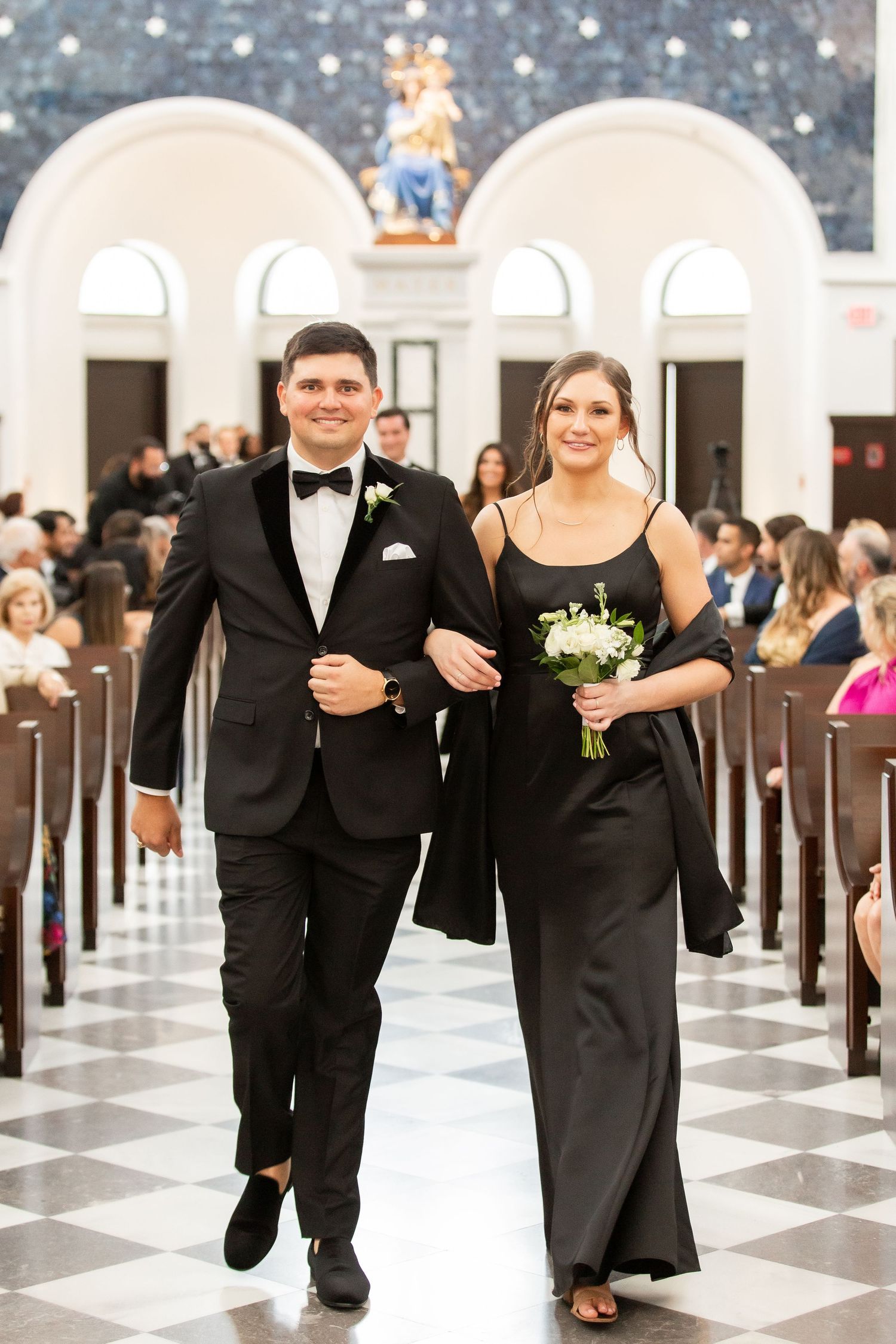 Formal wedding party members walking in pairs down an elegant church aisle with black and white floors.