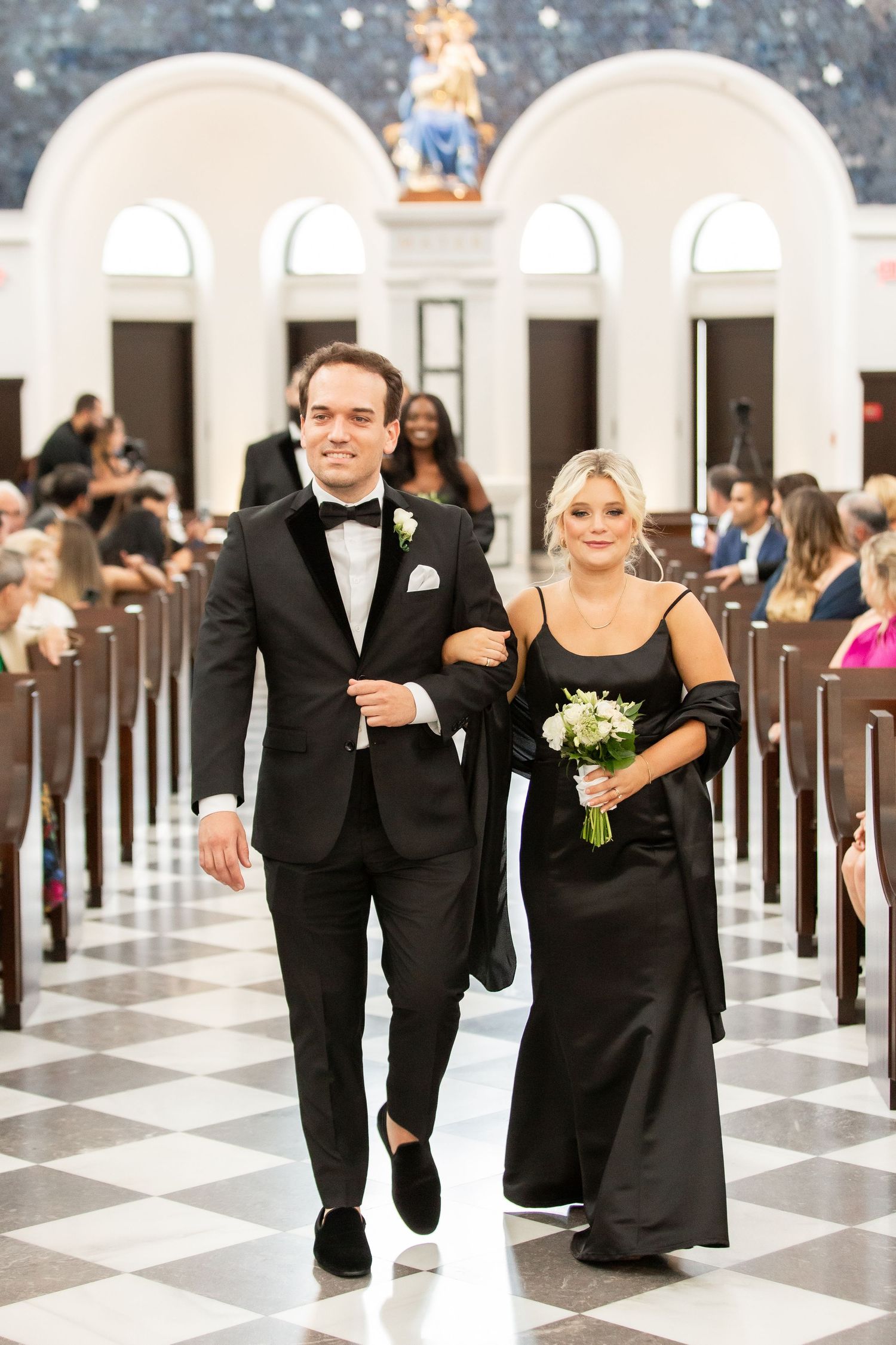 Couples in formal black attire walking together down a church aisle during a wedding ceremony.