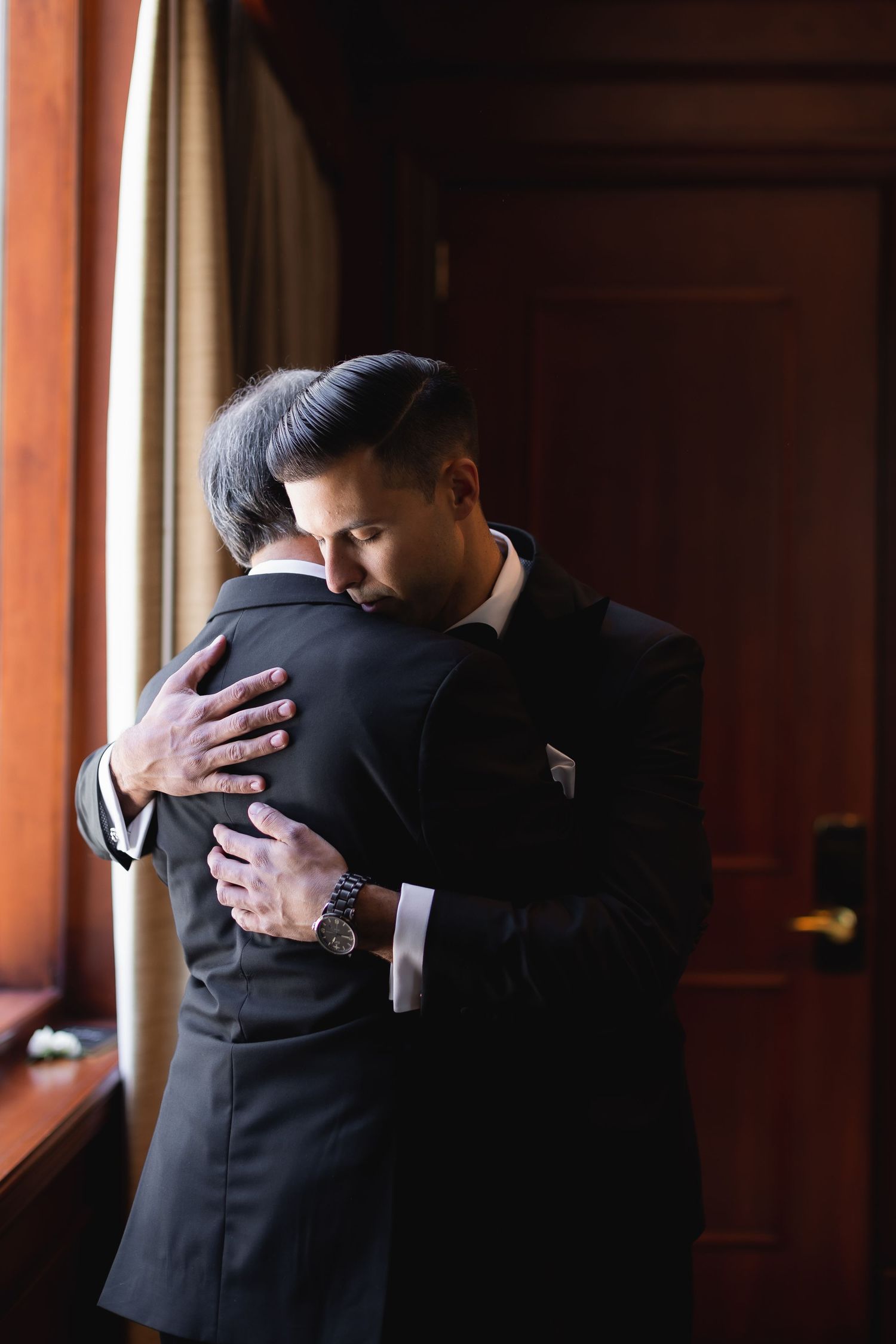 Two people in black suits embrace in a dramatic backlit scene.